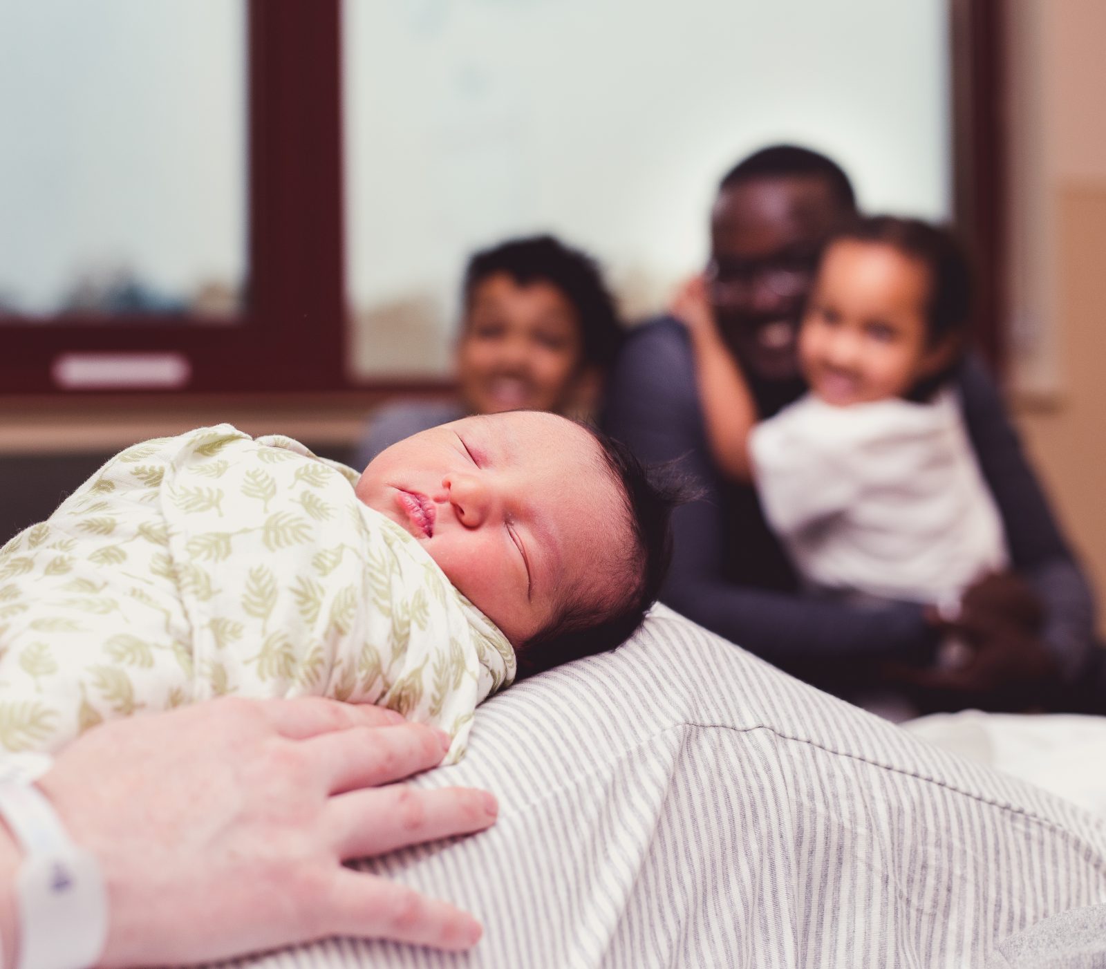 swaddled newborn sits on mom's knees with father and siblings in the background in the background
