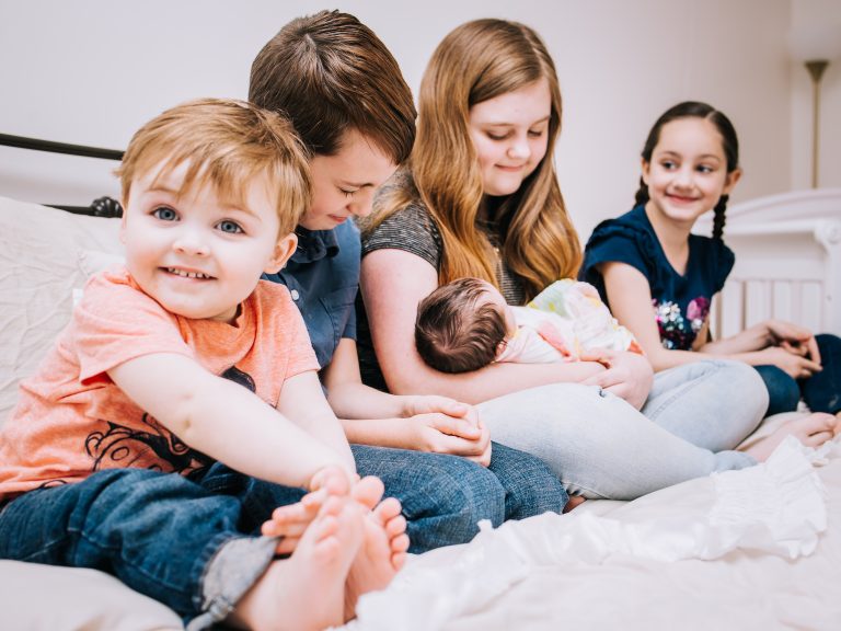 siblings sitting in a row smiling and holding new baby