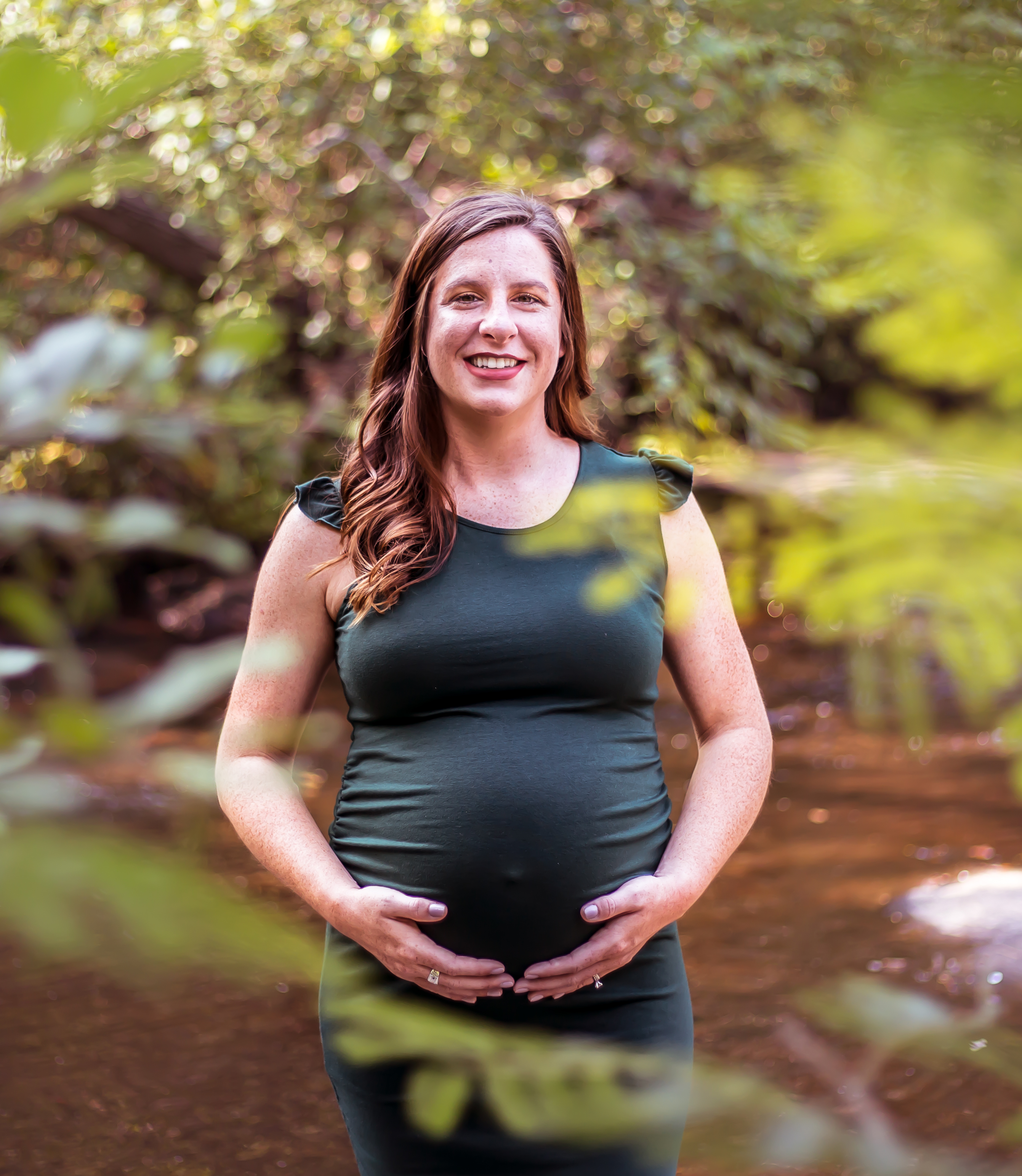 a pregnant woman stands with her hands on her belly, and there are tree leaves in the foreground