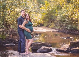 a pregnant woman and her husband stand on a rock by a stream