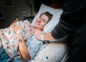 New mother holds her newborn just after birth. Her husband's hand rests on her sholder